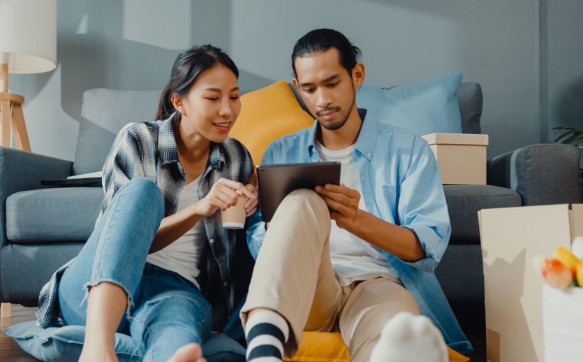 happy-asian-young-couple-man-woman-use-tablet-online-shopping-furniture-decorate-house-with-carton-packages-new-house
